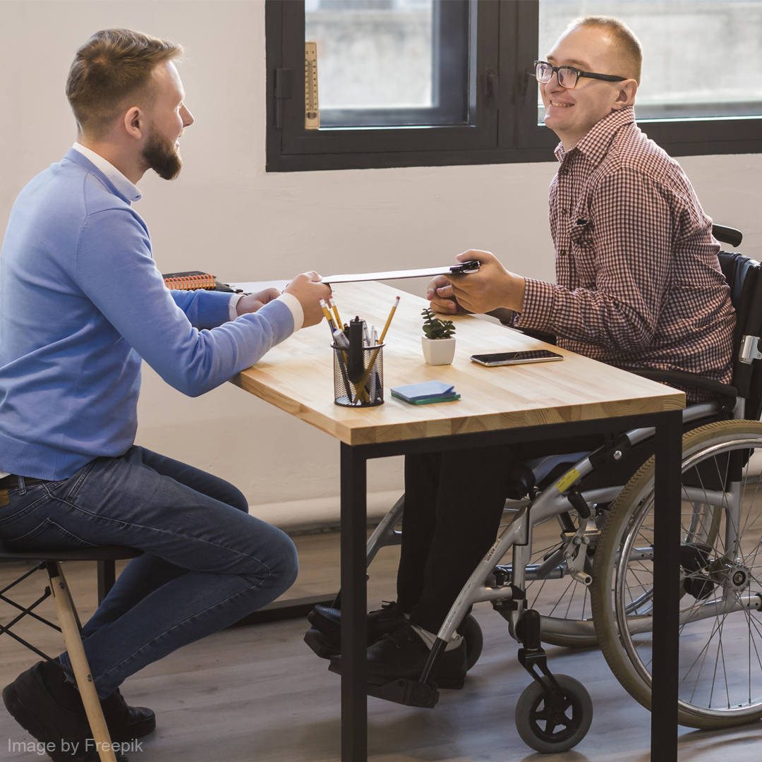 Ein Mitarbeiter sitzt mit einem Mann im Rollstuhl an einem Tisch und lachen gemeinsam.
